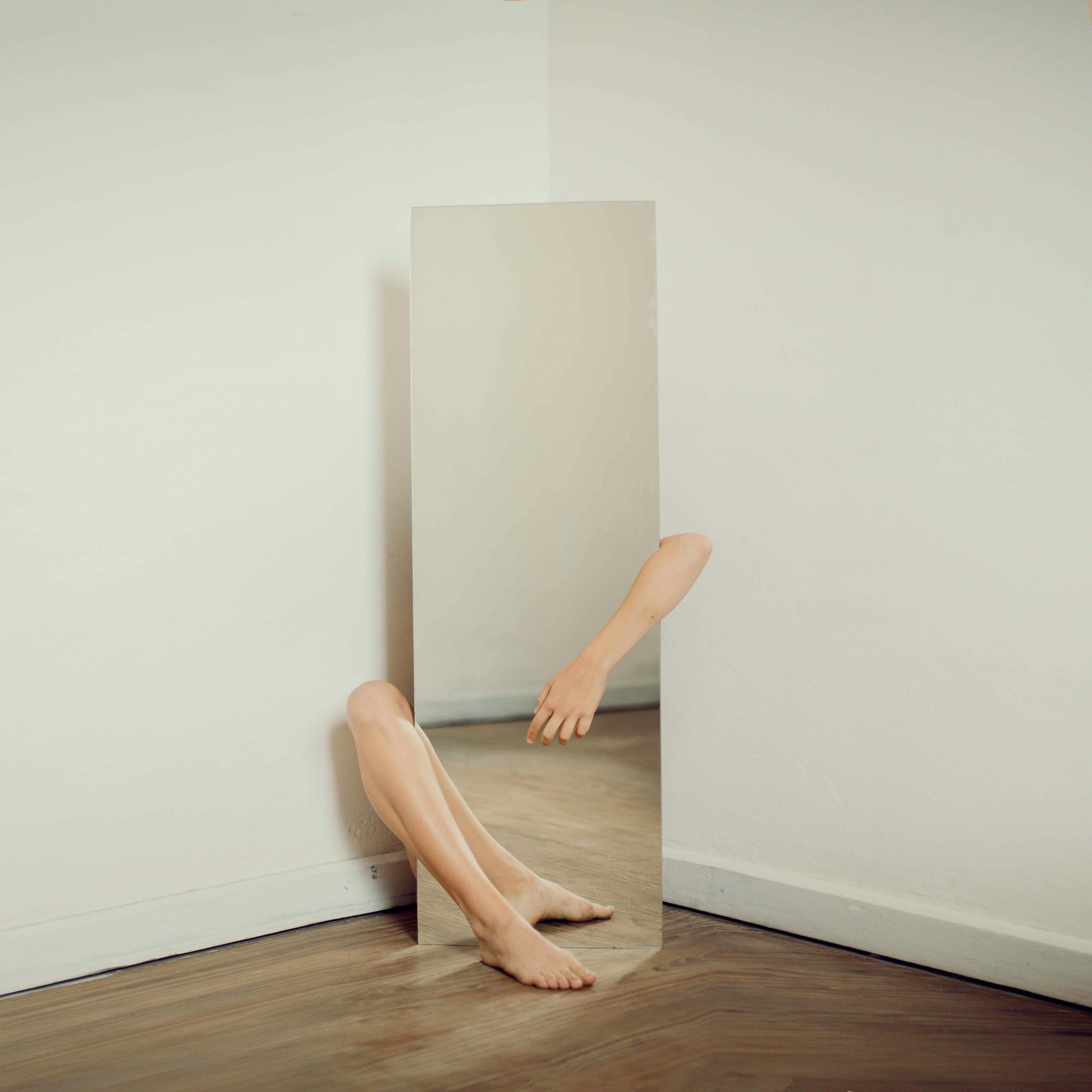 Surreal photo featuring a mirror reflecting an arm and leg on a wooden floor.